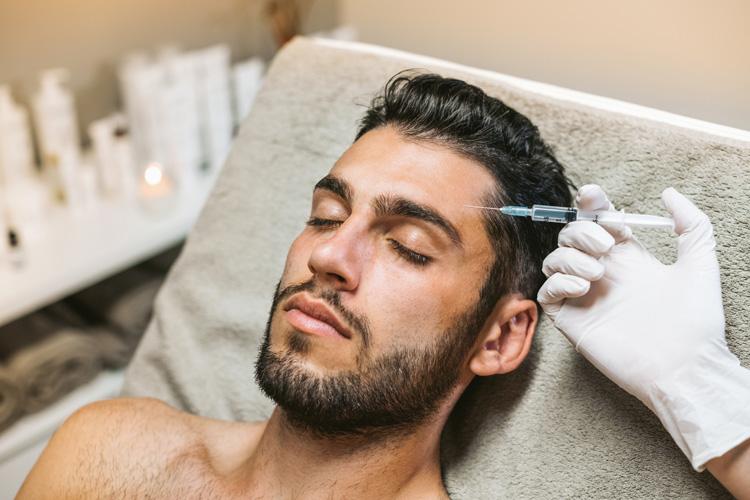 Man reclining in a treatment chair while an aesthetic practitioner positions a syringe near his temple for an injectable treatment.